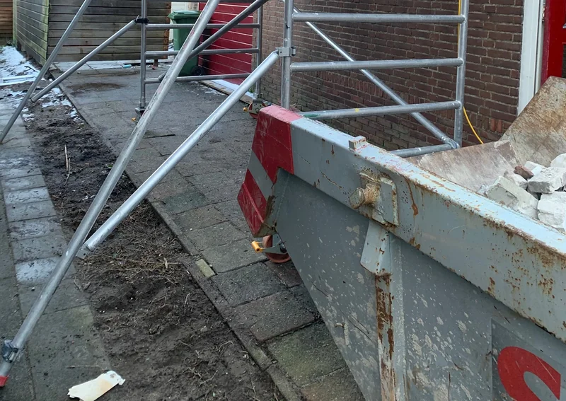 Een container met metselpuin naast een steiger en een gemetselde gevel.