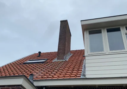 Gemetselde schoorsteen op een rood pannendak, vlak naast een dakkapel.