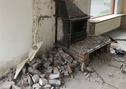 Gedeeltelijk gedemonteerd metselwerk rond een haard, met puin op de vloer.